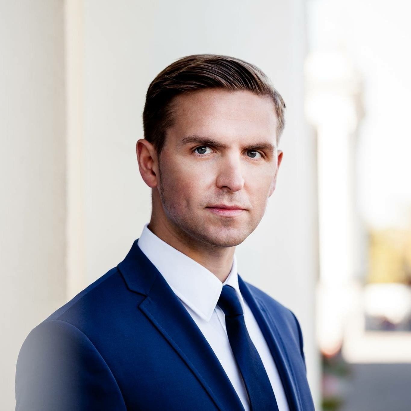 Man in blue suit with white shirt and tie, standing outdoors against a light background.