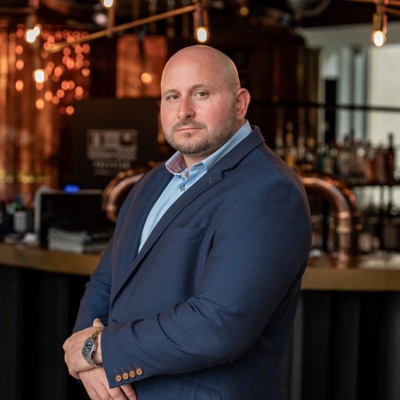 Man in a blue suit standing in a dimly lit bar with copper accents in the background.