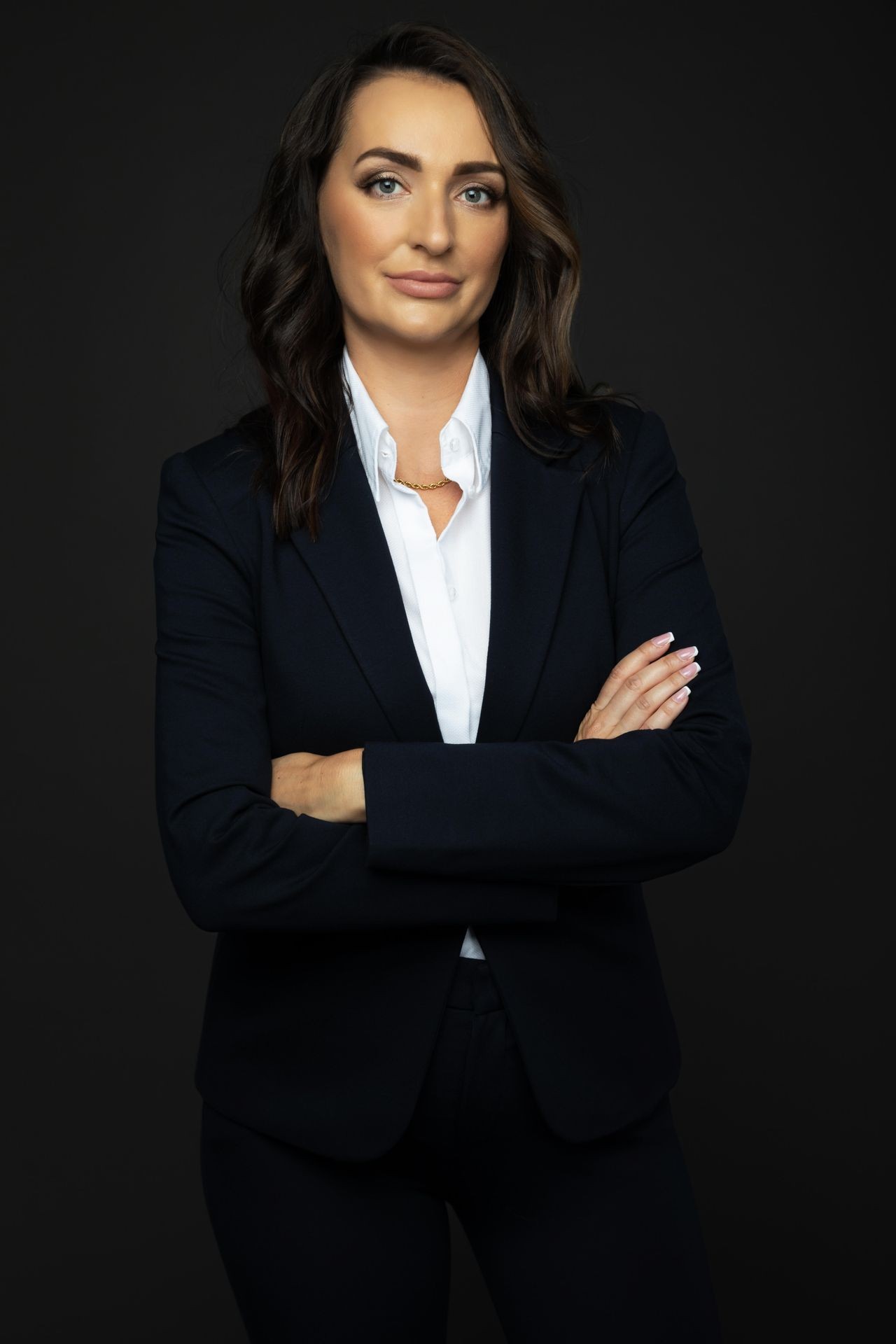 Individual posing with arms crossed, wearing a dark suit and white shirt against a black background.